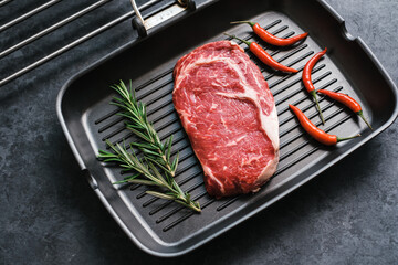 Raw rib eye beef steak on black grilling pan with chili pepper and rosemary, top view.