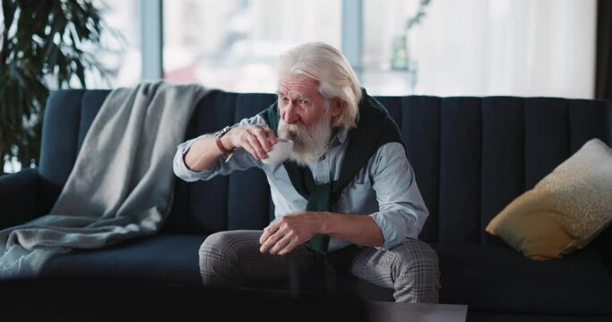 Mature pensioner bearded old caucasian man watching TV channel learning negative news drinking coffee and getting worried. Entertainment. Emotions.
