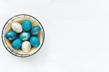 Easter basket with colored blue and gold eggs on a white wooden background with copy space. Horizontal easter banner with nest with eggs in minimal loft style. Modern ideas for Easter design.