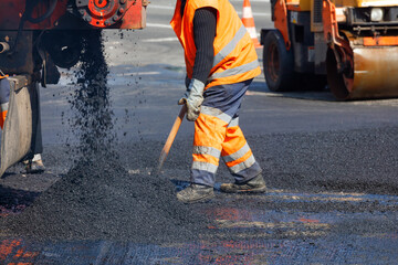 Fototapeta A road worker in orange overalls shovels fresh asphalt from a truck across a section of road being repaired. obraz