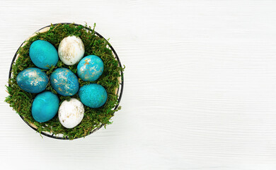 Easter basket with turquoise and gold eggs on a white wooden background top view. Easter banner with a basket of eggs with copy space. Basket with green moss and easter blue eggs for invitation design
