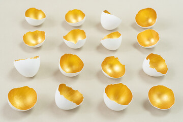 Eggshell, painted from the inside in gold color, lies in even rows on a beige background. Harmonious combination of gold, white and ecru colors. Concept for creative modern Easter greeting card.
