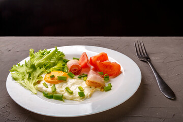 Scrambled eggs with bacon garnished with tomato and green onion on a gray concrete background near a fork.