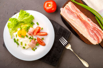 Scrambled eggs with bacon, garnished with tomato and green onion and on a gray concrete background on a wooden stand with a fork next to chopped bacon and tomato.