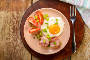 Scrambled eggs with bacon with tomato and green onions and on a wooden table in a plate on a stand with a fork.