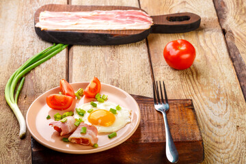 Scrambled eggs with bacon with tomato and onion and on a wooden table in a plate with a fork next to sliced bacon and tomatoes.