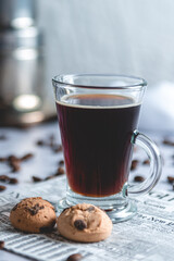 Taza de cafe con 2 galletitas sobre servilleta de papel con diseño de diario. Acompañan granos de cafe. 