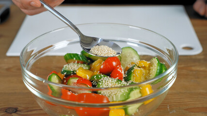Close up hand professional chef standing near kitchen table sprinkle sesame seeds on cooked vegetarian salad in glass bowl. Man preparing delicious healthy vegan salad dish for breakfast or dinner