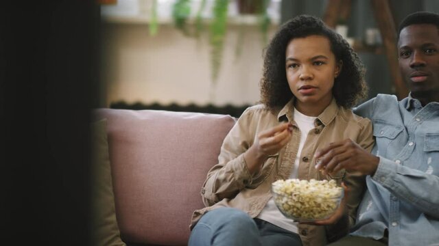Panning slow-motion waist-up shot of young afro couple sitting in front of TV in bright and cozy living room, eating popcorn and discussing film while watching