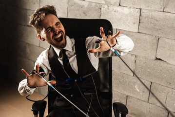 frightened, worried, pensive, man sitting on a chair, testing on a polygraph, wearing polygraph sensors, sitting on a gray brick wall background, wires held in his hands, holding hands, top view.