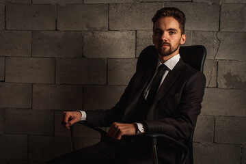businessman, serious and business man with a beard in a dark suit and white shirt sitting on a dark leather office chair on a gray background. low key. Selective focus