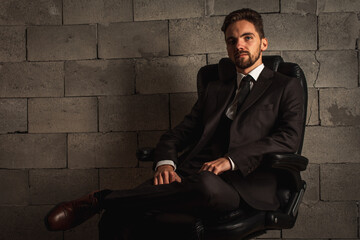 businessman, serious and business man with a beard in a dark suit and white shirt sitting on a dark leather office chair on a gray background. low key. Selective focus