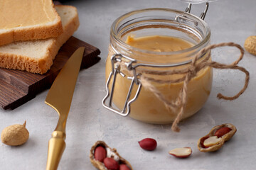 Glass jar with natural peanut butter, golden colored knife, peanuts and toast sandwiches on a light background. Vegetarian traditional breakfast close up.