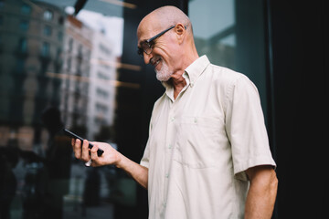 Cheerful male blogger 60 years old enjoying smartphone chatting during walk time at urban street, happy Caucaisan man on retirement using 4g wireless connection on mobile gadget for web phoning