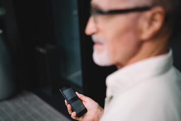 Blurred male blogger using cellphone device during daytime for travel walk at urban setting, unrecognizable aged man with smartphone technology thoughtful looking away standing at city street