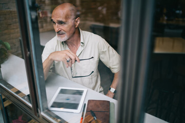 Fototapeta premium Window view of old Caucaisan journalist with optical glasses in hand thoughtful looking away, elderly male blogger with modern touch pad for browsing thinking about web information in cafe interior