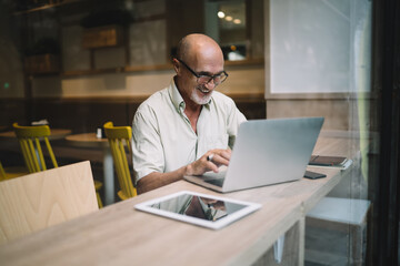Mature cheerful man in spectacles typing on laptop computer share content in blog browsing web pages and information, happy male freelancer doing remote job using netbook in coworking and smiling