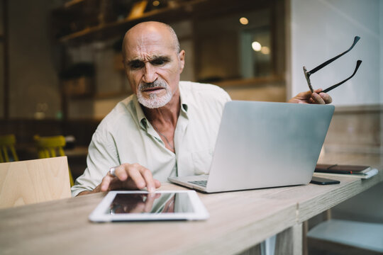 Puzzled Male Freelancer 60 Years Old Confused With Received Gadget Notification During Distance Working On Laptop, Skilled Aged Man Checking Email Message On Modern Digital Tablet Using 4g Internet