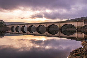 Long arched bridge crossing Ladybower reservoir reflected in Derwent Valley still deep river water with sunrise beautiful orange sky setting behind in the Peak District landscape