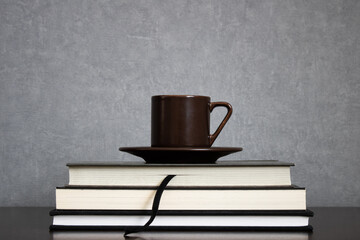 Fototapeta premium Brown cup of coffee with saucer on top of pile of books to read centered on a clean gray background. Empty space for text. Study, reading or preparing for exams concept