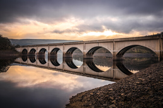 Long arched bridge crossing Ladybower reservoir reflected in Derwent Valley still deep river water with sunrise beautiful orange sky setting behind in the Peak District landscape