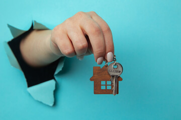 Woman's hand with house keys through a hole in blue paper close-up. House sale and rent concept.