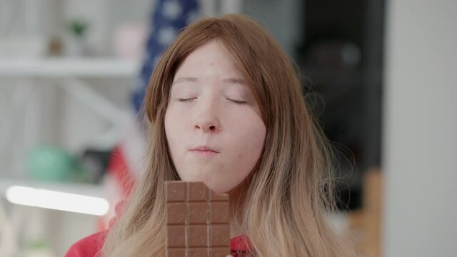 Close-up portrait of Asian girl with piercing in nose eating tasty sweet chocolate bar and smiling. Teenager with problematic skin enjoying delicious dessert at home indoors. Adolescence and lifestyle