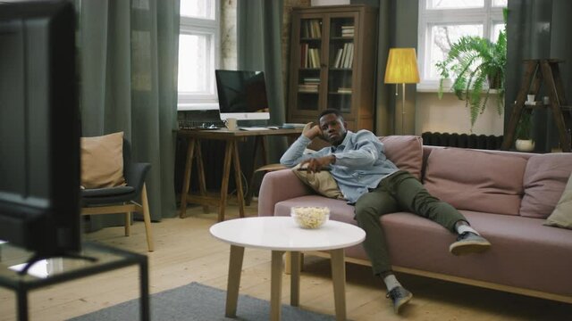 Panning slow-motion full shot of bored african american man lying on sofa in living room switching channels on tv with remote control, searching for program to watch