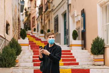 Man using mobile phone and wearing medical masks outdoors. Tourist in empty city street. Tourism during pandemic. Spain flag on steps. Travel bans countries set limits for travel face masks mandatory