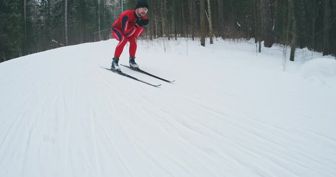 Cross country ski workout. Young man in red ski suit works out and glides downhill on the cross country ski track in a forest