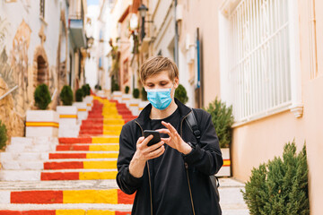 Man using mobile phone and wearing medical masks outdoors. Tourist in empty city street. Tourism during pandemic. Spain flag on steps. Travel bans countries set limits for travel face masks mandatory