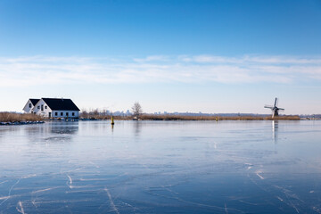 Fototapeta premium Winterlandscape with ice on the Dieperpoel (Kagerplassen) in front of a windmill (de Kok) and house on the Kogjespolder in the south-holland municipality of Teylingen (Sassenheim) in the Netherlands.