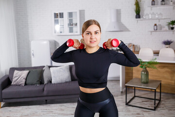 Fototapeta na wymiar Young pretty woman in a black top with dumbbells in her hands doing sports at home, fitness at home during quarantine. Healthy lifestyle