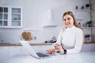 Beautiful young woman studying at home on a laptop. A girl with long blond hair in a sweater drinks tea sits in the kitchen. Work remotely from home