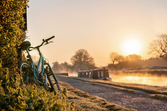 Canal boat with mountain bike left leaning against hedge row early morning sunrise dawn with golden light in sky on the River Trent and mist rising in Nottingham biker on ride out