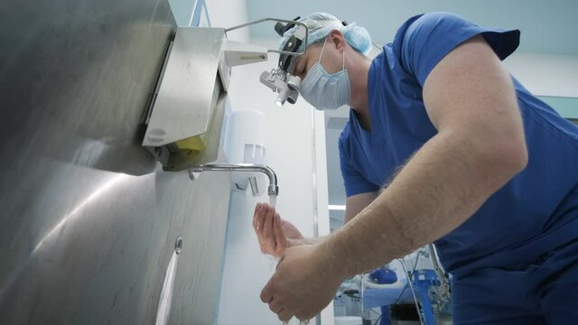 The surgeon prepares for the operation, washes and sterilizes his hands in the modern operating room. Doctor is equipped in mask, medical headlamp, surgical loupes