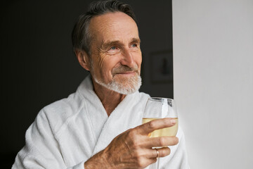 Close up of smiling mature man wearing white bathrobe holding wineglass looking away