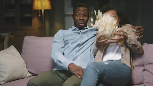 Zoom-in slow-motion POV of young afro couple watching breathtaking horror movie sitting on sofa in living room. Girl throwing bowl with popcorn from fear and they starting laughing together