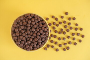 Chocolate cereal corn balls in a yellow bowl scattered on a yellow background. Top view. Copy, empty space for text