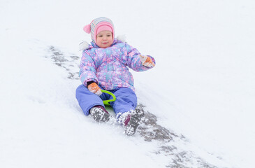 A little pretty girl in a purple overalls slides down an ice slide, winter children's games, a...