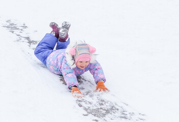 A little pretty girl in a winter overalls is riding on ice slides in winter, a child is driving...