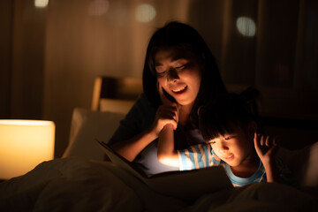 Little girl looking and reading a homework on the book at home with her mother, She has her head on her hand and her mother is explaining something, night shot with cozy warm light, mother teaching