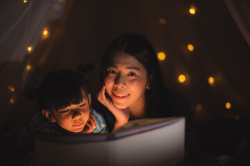 Little girl looking and reading a homework on the book at home with her mother, She has her head on her hand and her mother is explaining something, night shot with cozy warm light, mother teaching