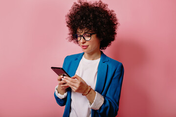 Close up studio shot of successful young lady with curls wearing blue jacket and glasses using...