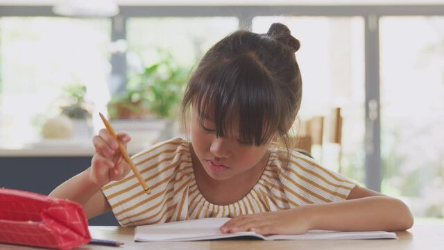Front view of young Asian girl home schooling working at table in kitchen writing in book during lockdown - shot in slow motion