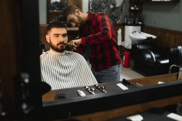 Fototapeta na wymiar Handsome bearded man is smiling while having his hair cut by hairdresser at the barbershop