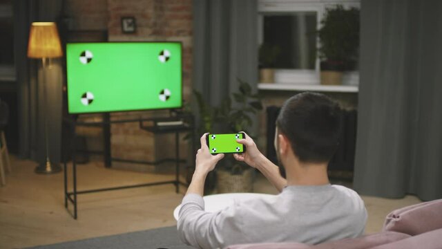 Rear-view Slow-motion Medium Close Up Of Male Football Fan Using Smartphone While Watching Football Match On TV