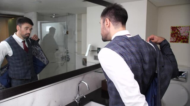 Handsome brunette man standing in front of bathroom mirror and putting on suit jacket. Confident bearded male looking at his appearance in mirror before important business meeting