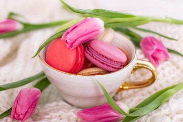 Sweet dessert, macaroons in golden mug and pink tulips on white wool background