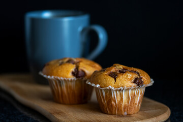 Freshly baked chocolate chip muffins and a cup of coffee on a wooden platter with a black background